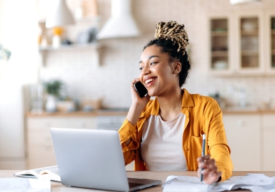 Woman with dark hair in braids and yellow jacket talking on her cellphone in front of a laptop taking notes