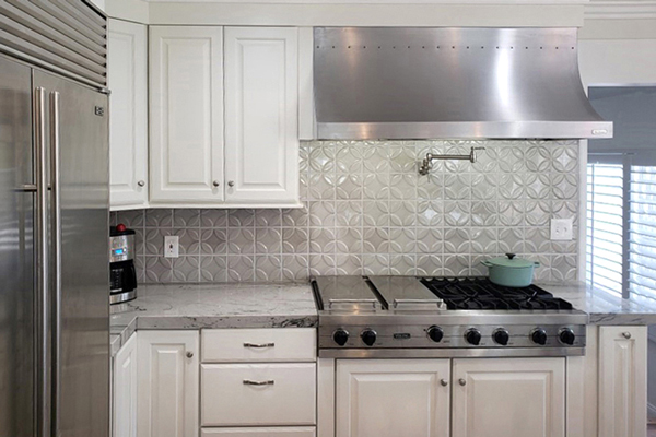 Stainless steel cooktop with matching range hood, wall faucet, grey tile backsplash and white cabinets