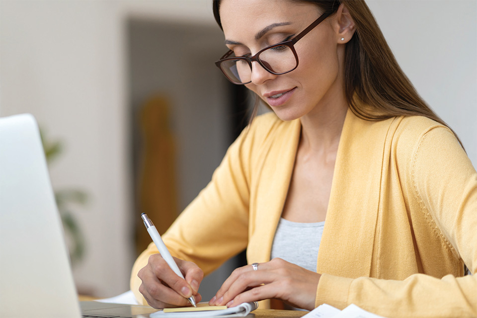 Brunette with dark rimmed glasses on and a yellow sweater writing down notes