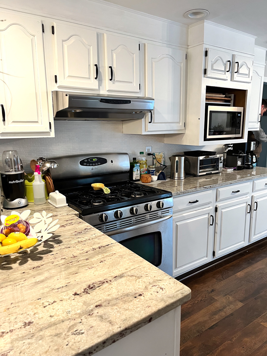 Client's before kitchen photo with partial overlay cabinets with white arched raised panel doors, exposed black hinges, soffits over the wall cabinets, and an awkward layout