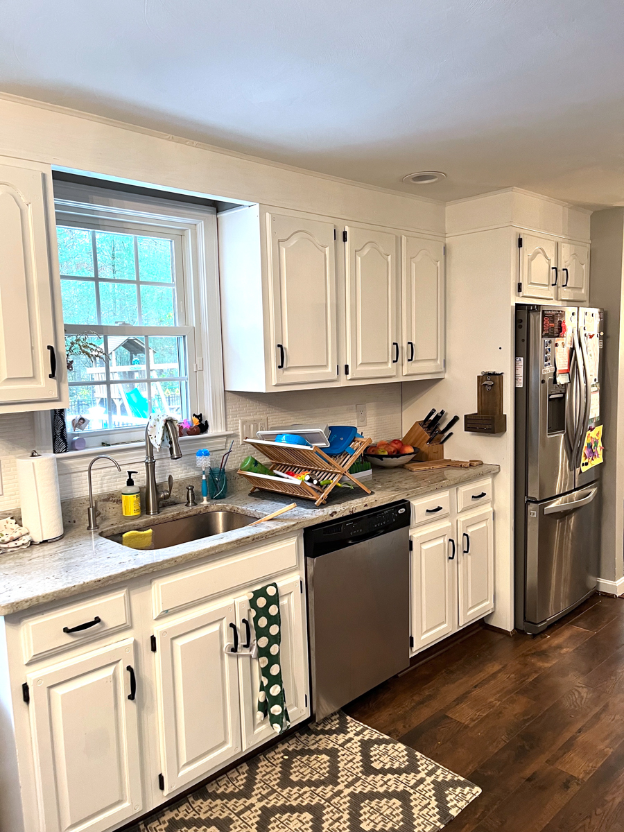 Client's before kitchen photo with partial overlay cabinets with white arched raised panel doors, exposed black hinges, soffits over the wall cabinets, and an awkward layout