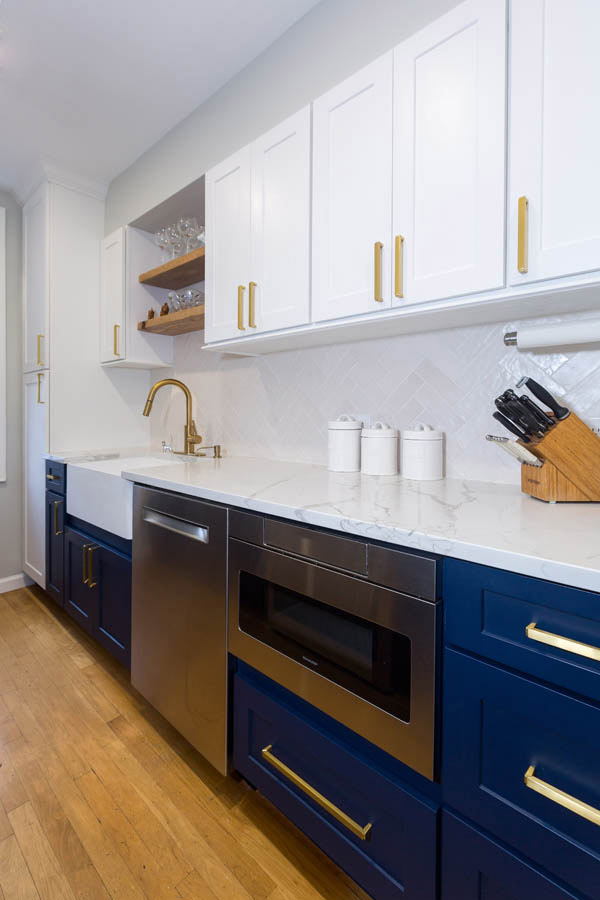 white kitchen with blue base cabinets, microwave cabinet next to stainless dishwasher and wood floating shelves above the sink