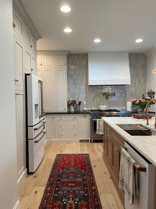 Cream colored inset shaker kitchen cabinets with warm wood-toned island, tan quartz countertops and textured glazed tile backsplash behind a tapered kitchen hood