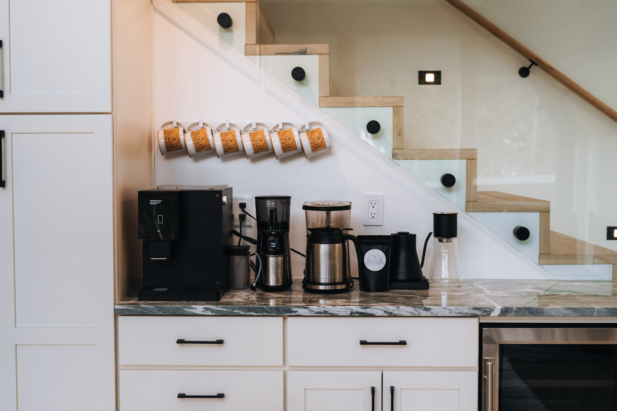 White shaker coffee bar cabinets next to a glass staircase railing with black cabinet pulls and coffee cups above