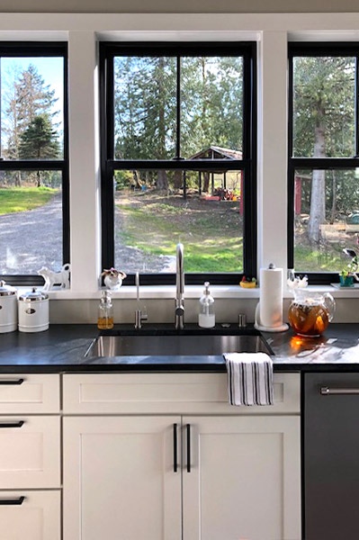 kitchen sink underneath three open windows with black trim looking out on a small farm, white shaker cabinets with black countertops, wash rag draped over the edge