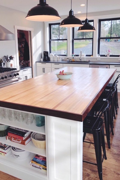 farmhouse style kitchen with butcher block countertops on the island under three black pendant lights, seating for four and open shelving in white Shaker cabinets