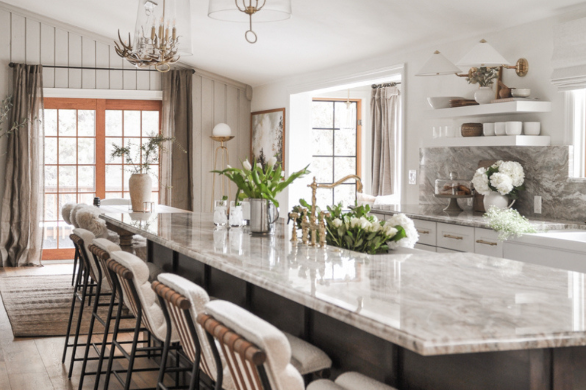 Luxe white stacked shaker kitchen cabinets, brass hardware, bronze metal kitchen hood, cream and brown marble countertops plus matching slab backsplash, and a large dark brown stained furniture-style island