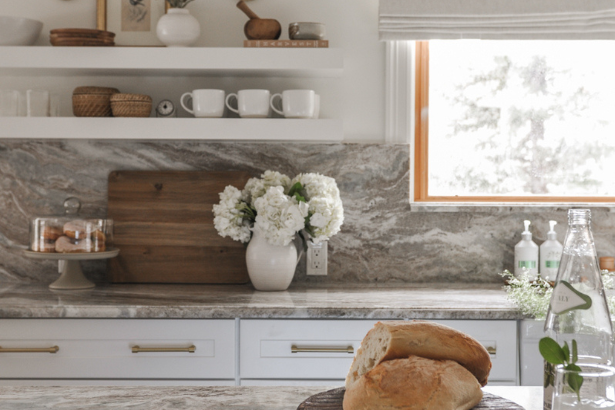 White shaker kitchen cabinets with cream and brown marble countertop and matching backsplash with floating shelves, sconces, and brass hardware