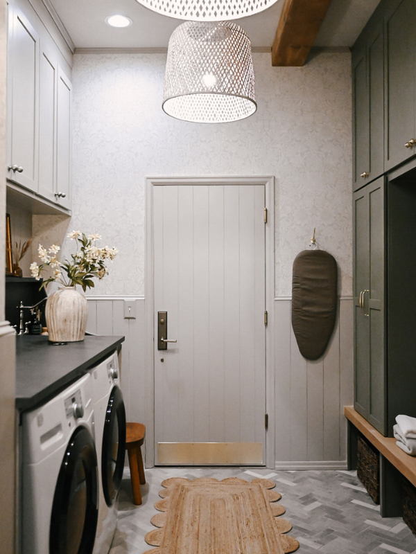 Large laundry room with washer/dryer on left with taupe shaker cabinets above and decorative black countertop across with olive green mudroom lockers and white oak bench on the right