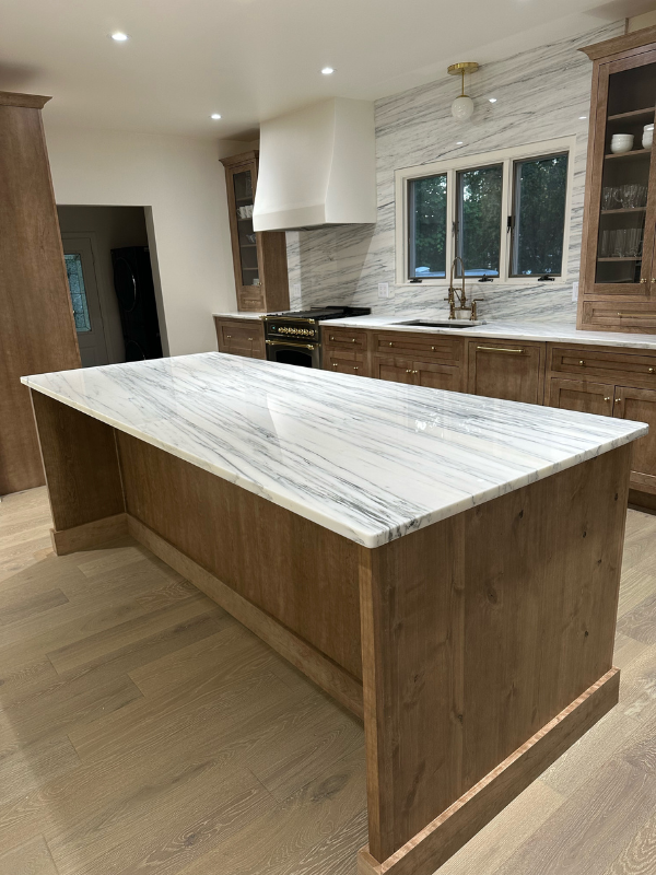 Medium-brown inset cabinets with furniture-style tall glass wall cabinets sitting on a white stone countertop with an artisan white kitchen hood