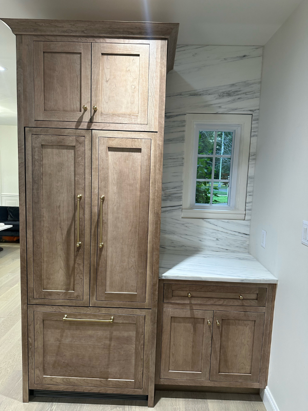 Refrigerator wall with a base cabinet on the right in medium-brown inset cabinets with gold hardware and custom panels covering and enclosing the refrigerator