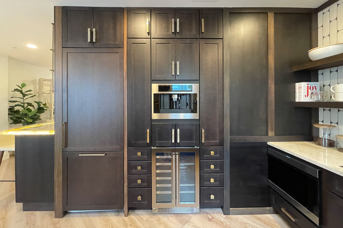 Small and chic U-shaped kitchen in dark brown shaker cabinets with wood floating shelves, white funky tile backsplash, and light brown quartz countertop with waterfall edge detail