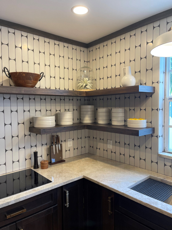 Small and chic U-shaped kitchen in dark brown shaker cabinets with wood floating shelves, white funky tile backsplash, and light brown quartz countertop with waterfall edge detail