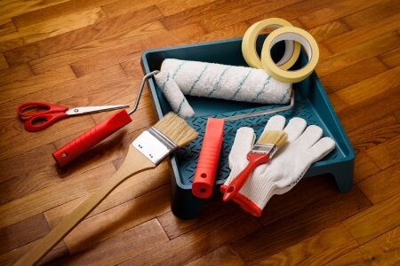 Paint pan on a wood floor with brushes, rollers, tape, gloves and scissors laying inside. 