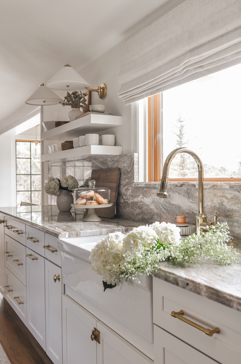 White shaker kitchen cabinets with cream and brown marble countertop and matching backsplash with floating shelves, sconces, and brass hardware