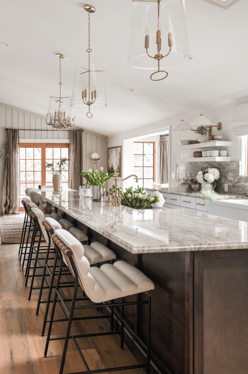 Luxe white stacked shaker kitchen cabinets, brass hardware, bronze metal kitchen hood, cream and brown marble countertops plus matching slab backsplash, and a large dark brown stained furniture-style island