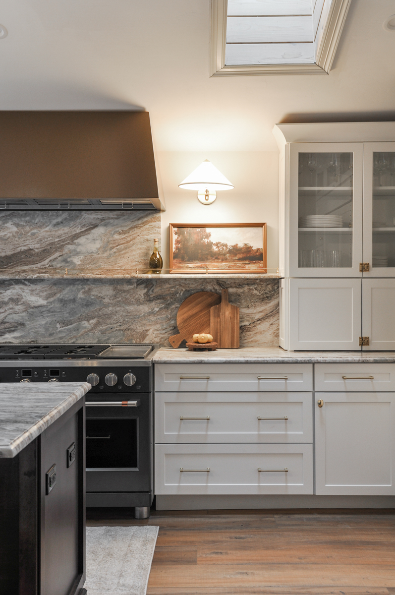 Stacked white shaker kitchen cabinets with glass on top, brass hardware, a bronze metal kitchen hood, and cream and brown countertops and backsplash with a stone ledge