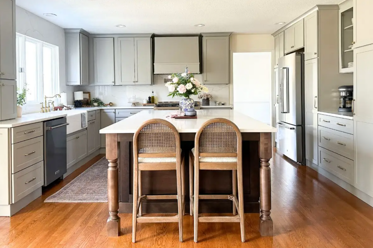 Light gray shaker L-shaped kitchen with matching wood hood, dark brown island cabinets and decorative legs, and white quartz countertops and matching slab backsplash