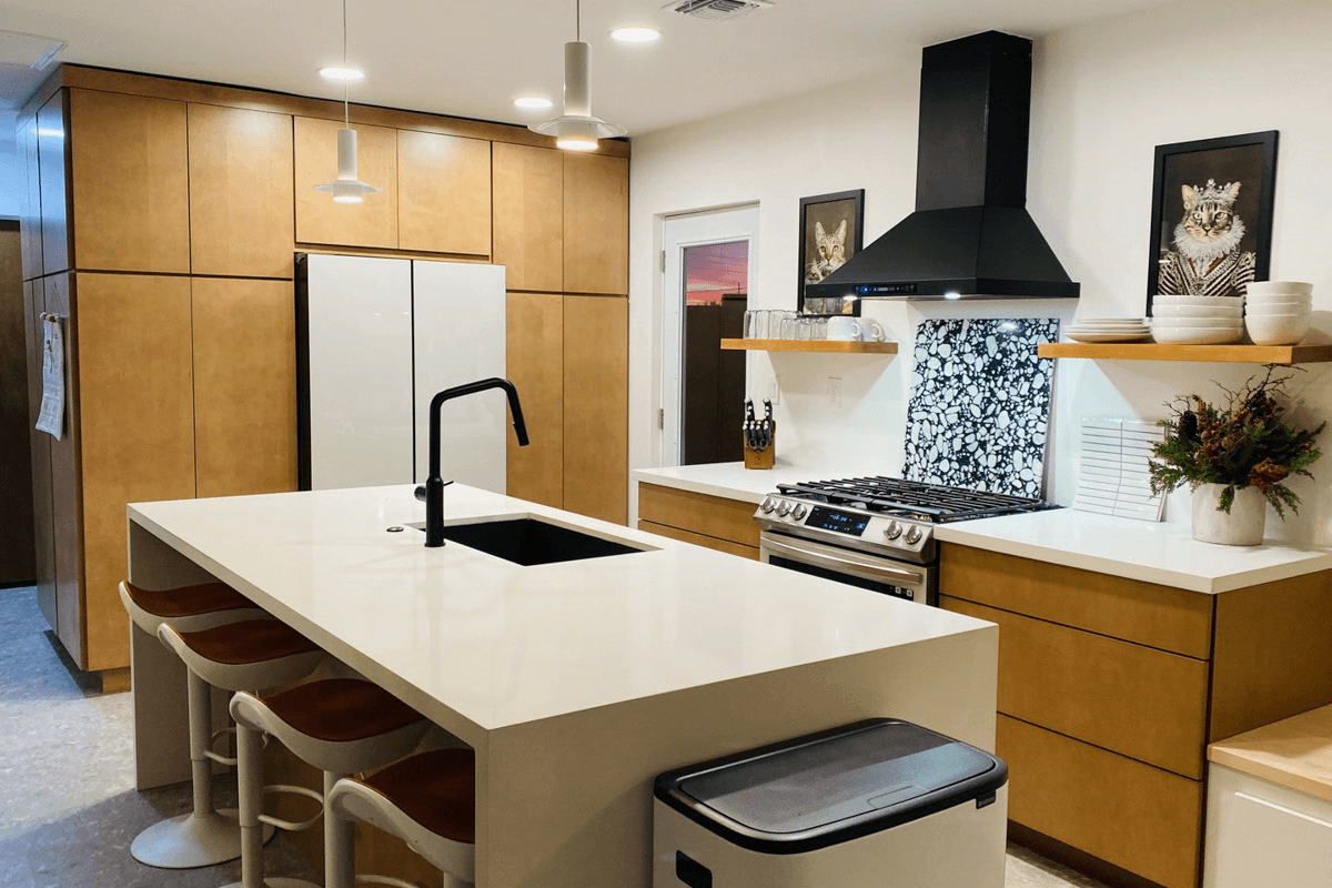 Mid-Century Modern L-shaped warm full overlay slab door kitchen design with cream quartz waterfall countertops, matching wood floating shelves and a black metal kitchen hood. 