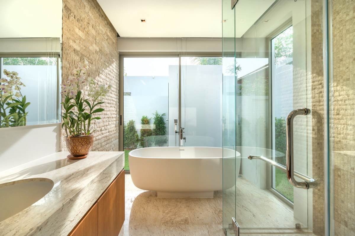Large organic bathroom with wood vanity cabinet and concrete countertop with freestanding tub in front of a large window next to focal wall in natural stone