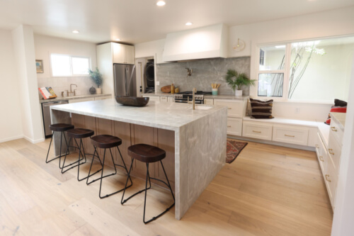 U-shaped shaker kitchen with cream cabinets in Mid-Century Modern style with large plaster range hood and medium wood-toned island with gray waterfall countertops