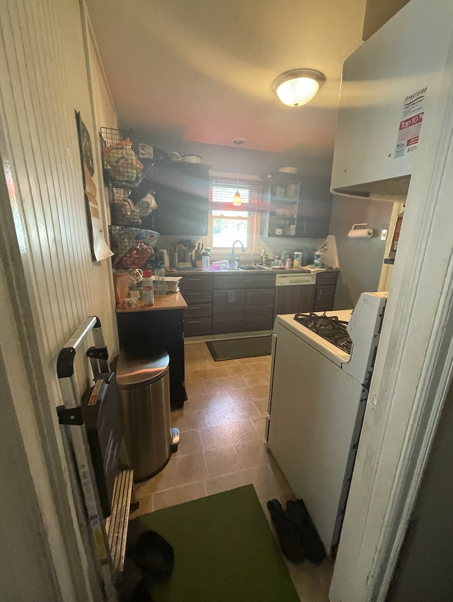 Over-crowded galley kitchen in dark and dingy finishes cluttered with household items 