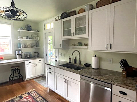 White shaker kitchen with dark countertops, floating wood shelves, and a built-in desk with seating