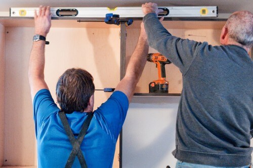 Two contractors installing wall cabinets and double-checking accuracy with a large level across the top