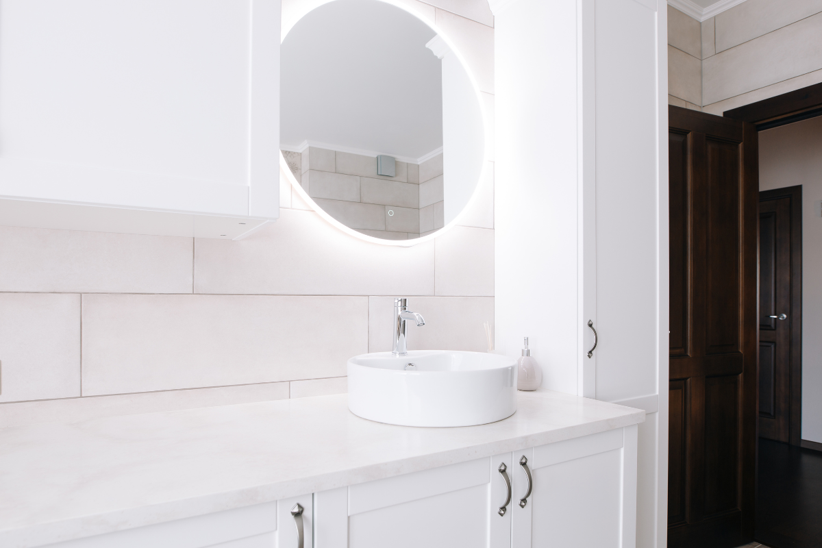 White shaker bathroom vanity cabinet with satin nickel hardware and circular mirror lit from behind