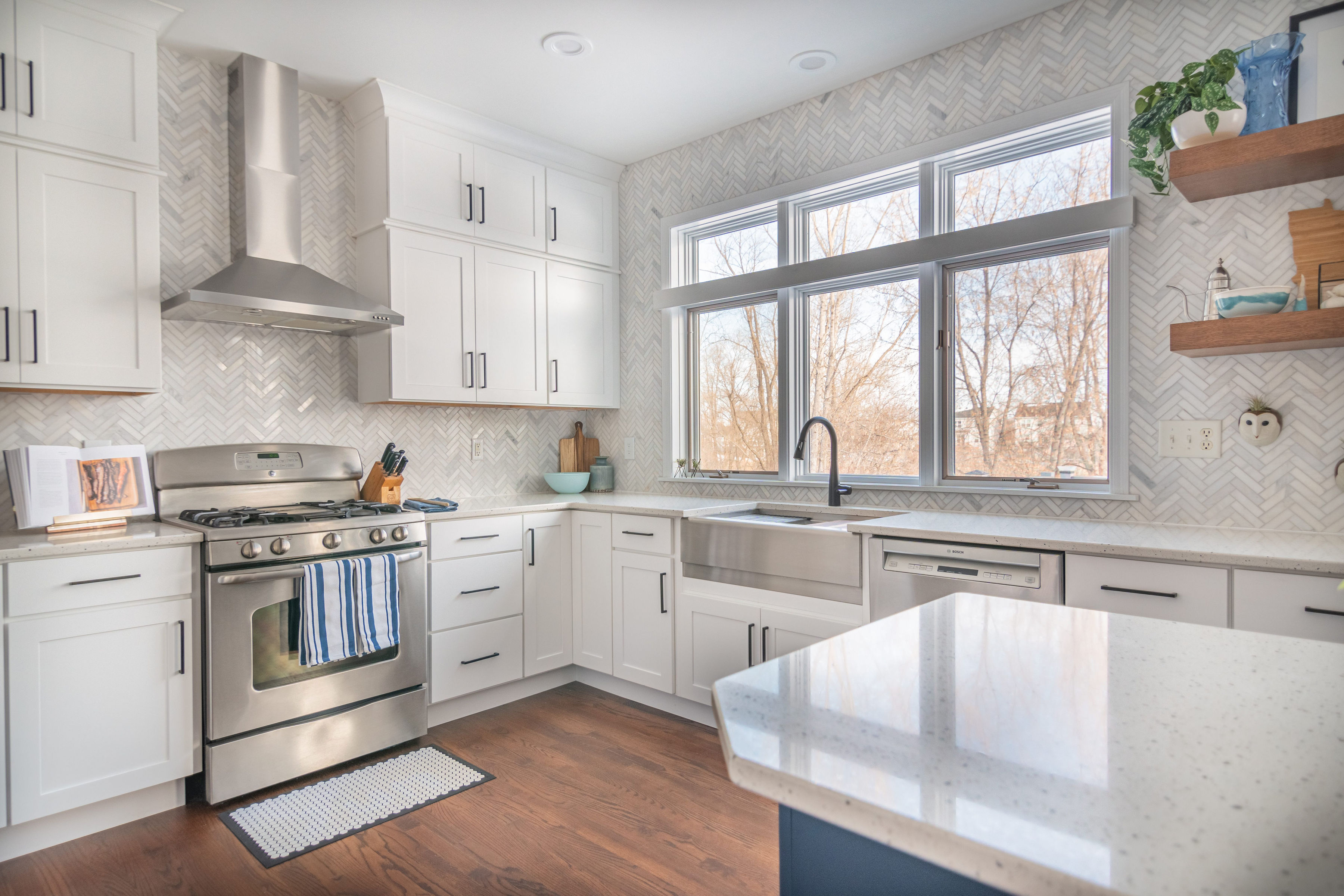 CliqStudios project: Stacked white shaker kitchen cabinets with a large window over the sink, marble herringbone tile, and warm wood floating shelves