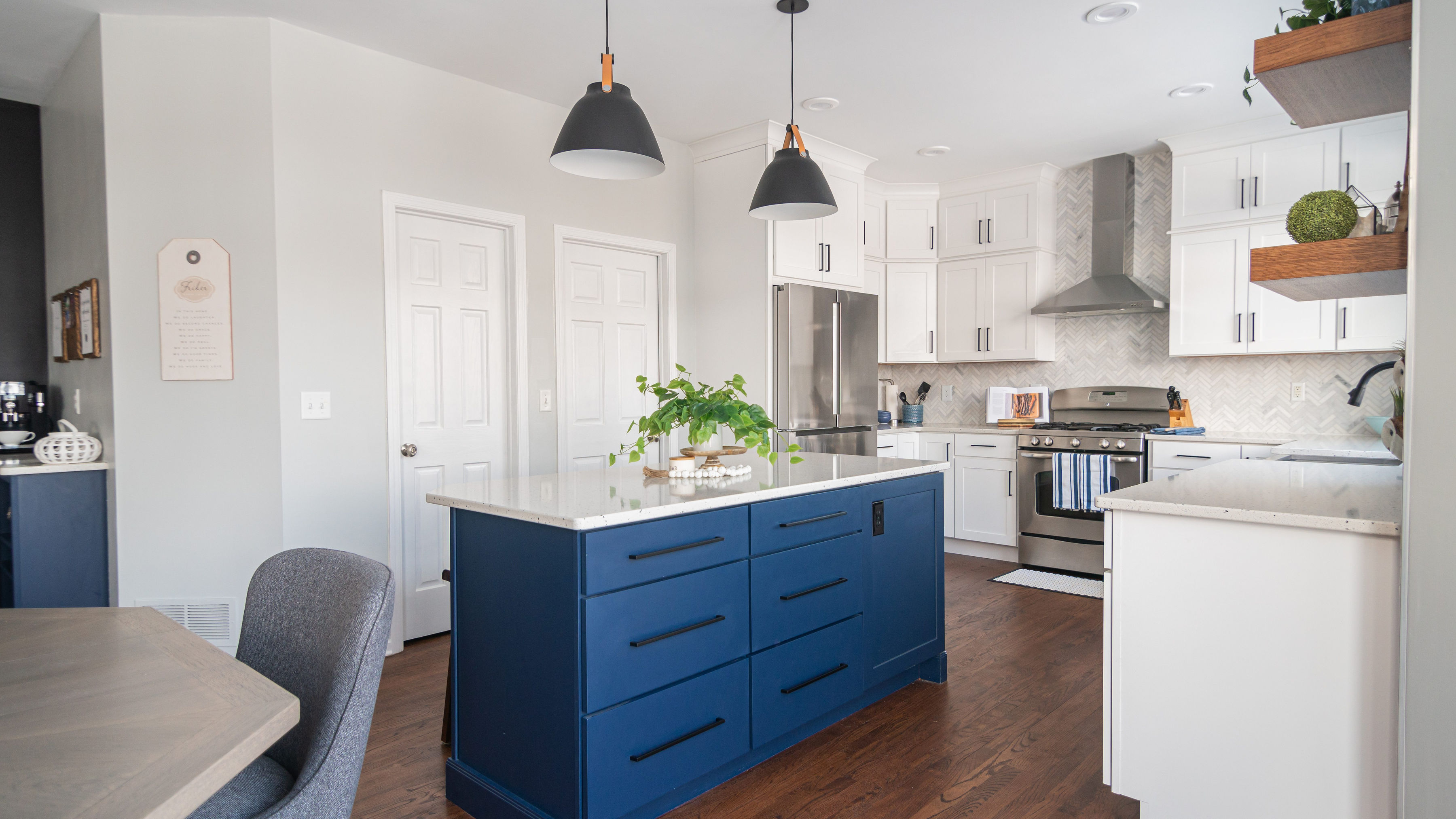 CliqStudios project: Stacked white shaker kitchen cabinets with blue shaker island, marble herringbone tile, and warm wood floating shelves