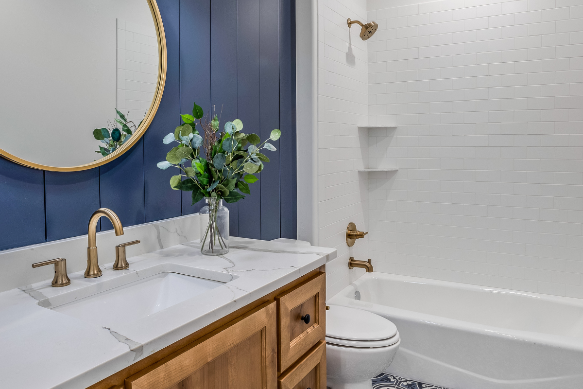 Light wood shaker vanity cabinet with marble countertop next to bright blue wall paneling and a matching round wood mirror