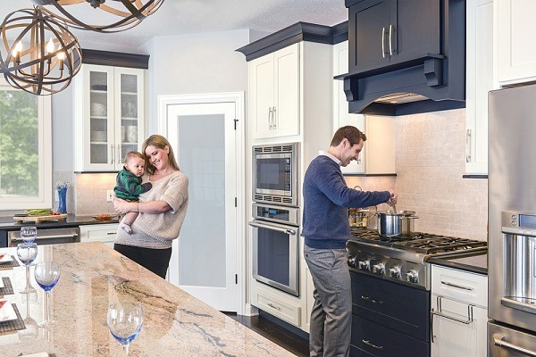 Homeowners Steve (at range) and Katie Delchin with baby in their kitchen,which features black and white cabinets, a center island and a professional range