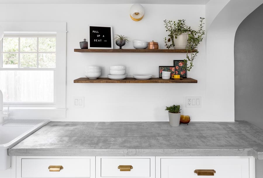 Open wood kitchen shelving above concrete countertops and white inset cabinets with gold hardware