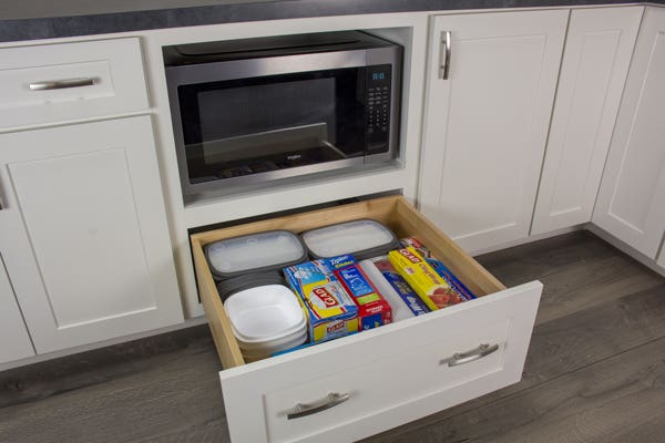 Open white drawer under a microwave shelf filled with food storage supplies