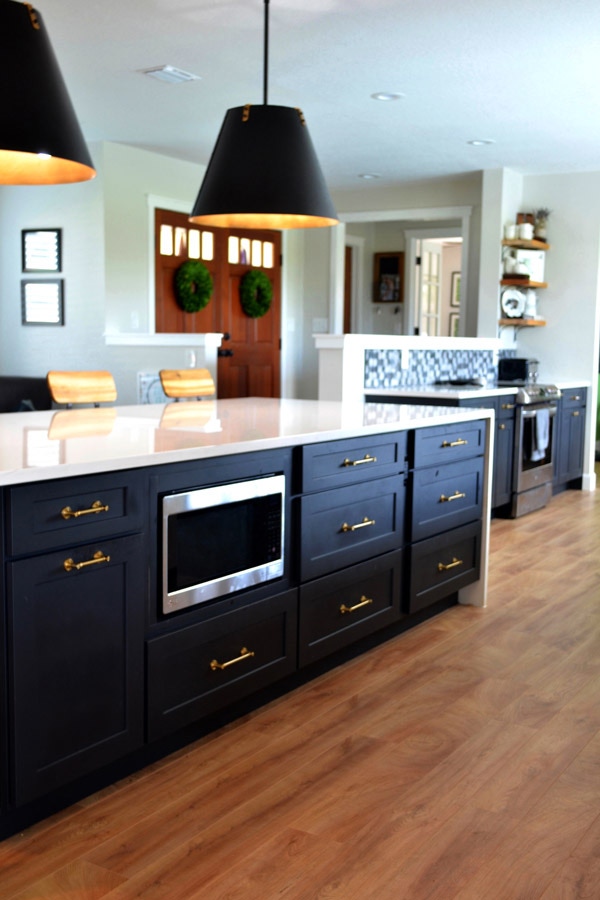 CliqStudios Shaker cabinets in Black used in a kitchen island with a white waterfall countertop. A base microwave sits in the center of the island, and pendant lighting with black shades hang above.