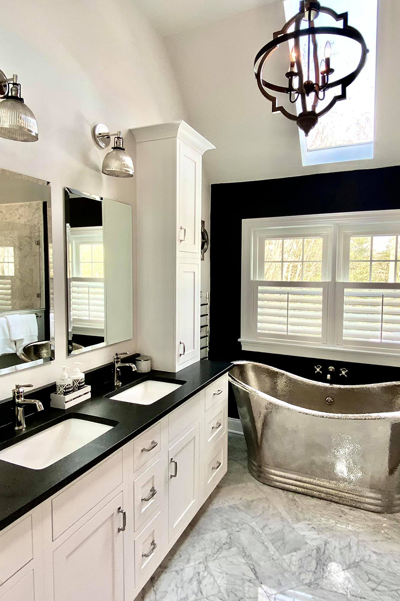 White inset shaker vanity with wall cabinet towers, black quartz countertops, a large metal soaking tub, and vaulted ceilings