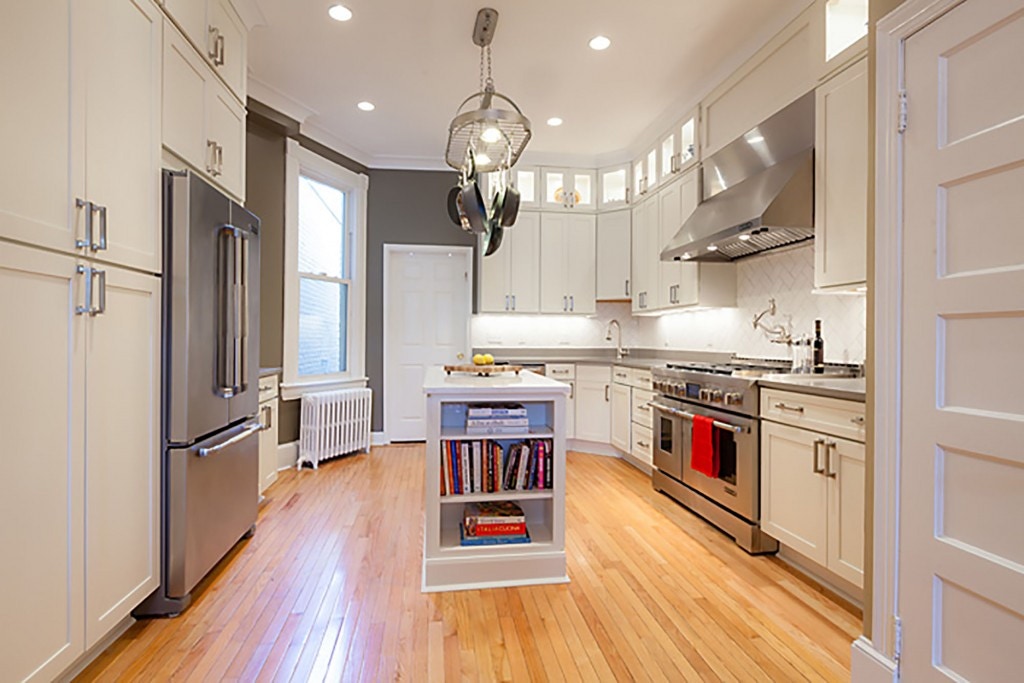 long view of remodeled kitchen in Washington DC townhouse, with refinished oak floors, hot water radiator, double refrigerators, white cabinets, bookshelf on end of center island and professional range and hood