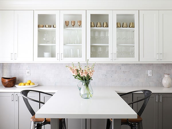 Two-toned cabinets in Shaker White and Medium Gray, with a white peninsula dining area featuring a vase of flowers and glass cabinet doors showing clear glasses and bronze cups