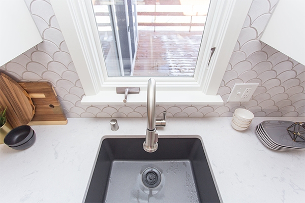 Downward facing view of a gray undermount sink with gooseneck faucet, a white arched tile backsplash and a deck view out of the window.