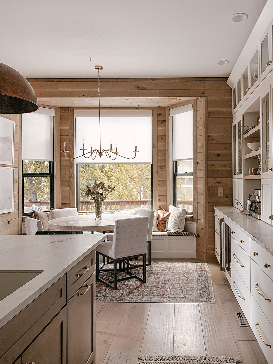 CliqStudios project: Light wood paneled breakfast nook with large window tucked in next to medium brown stained island and white shaker buffet kitchen cabinets