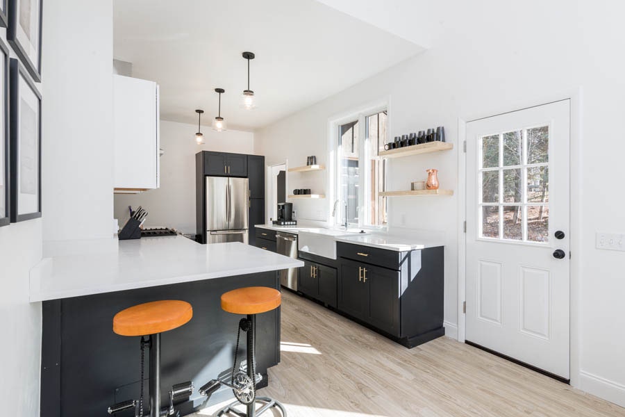 Brightly lit white galley kitchen with black base cabinets, white apron sink under a window with floating wood shelves on both sides