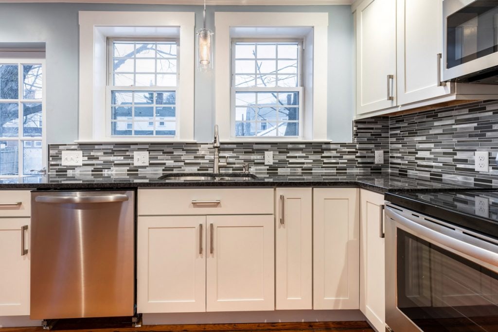 Kitchen sink facing two windows with white Shaker cabinets and black and white backsplash