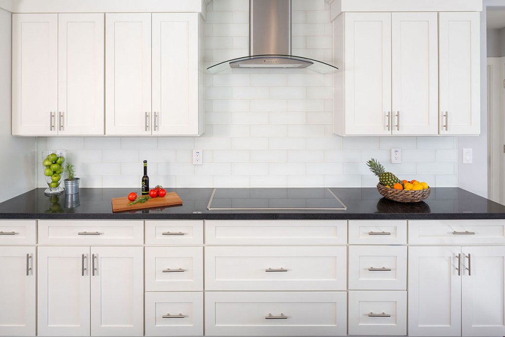 White shaker kitchen cabinets on a cooktop wall with black granite countertops and a stainless steel kitchen hood