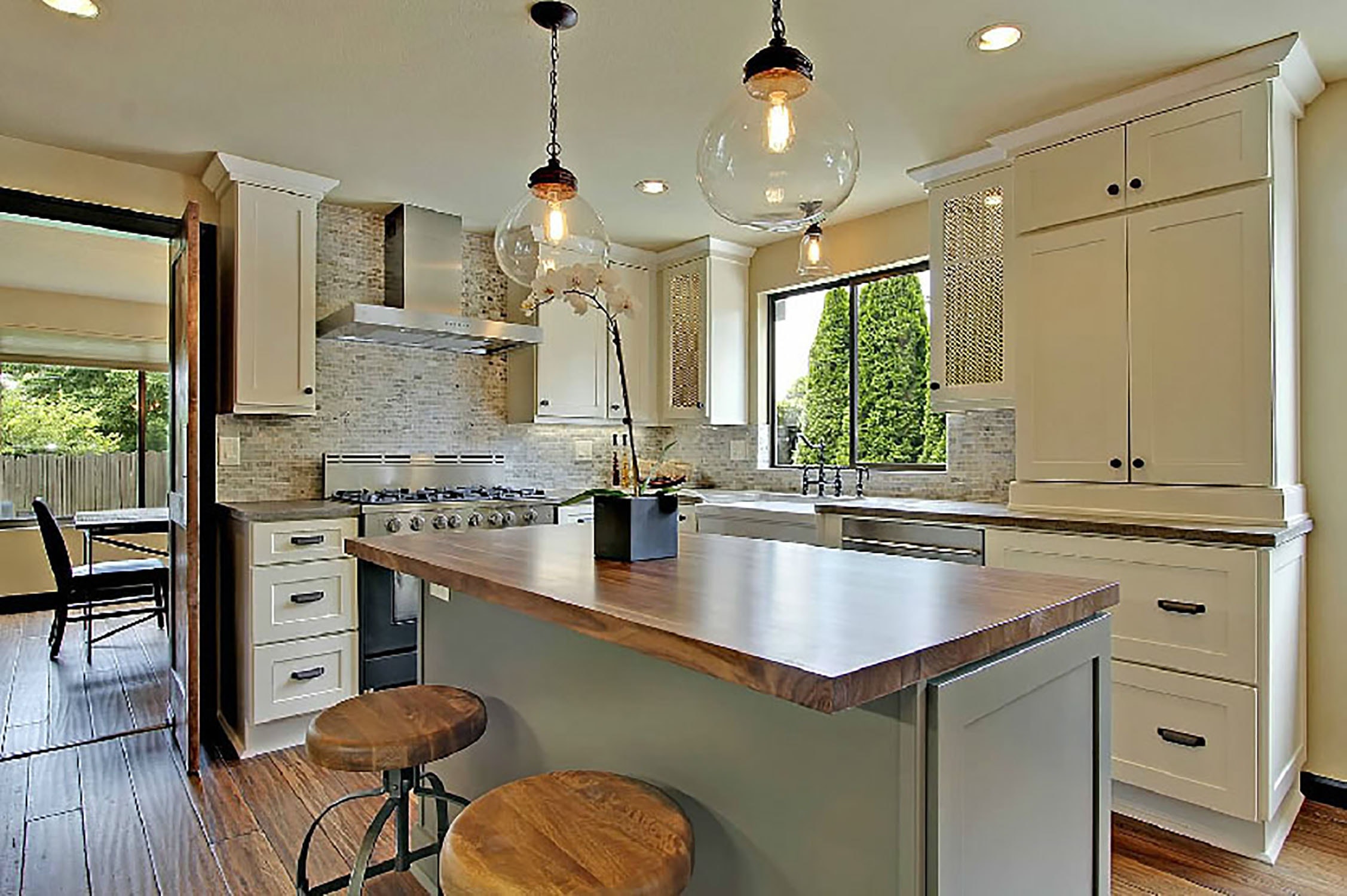 kitchen has white shaker cabinets, wood countertops, bar stools and floor, and natural stone backsplash