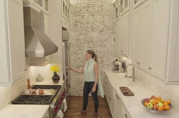 woman in New York kitchen with whitewashed brick walls, white shaker cabinets, farmhouse sink and glass door cabinets