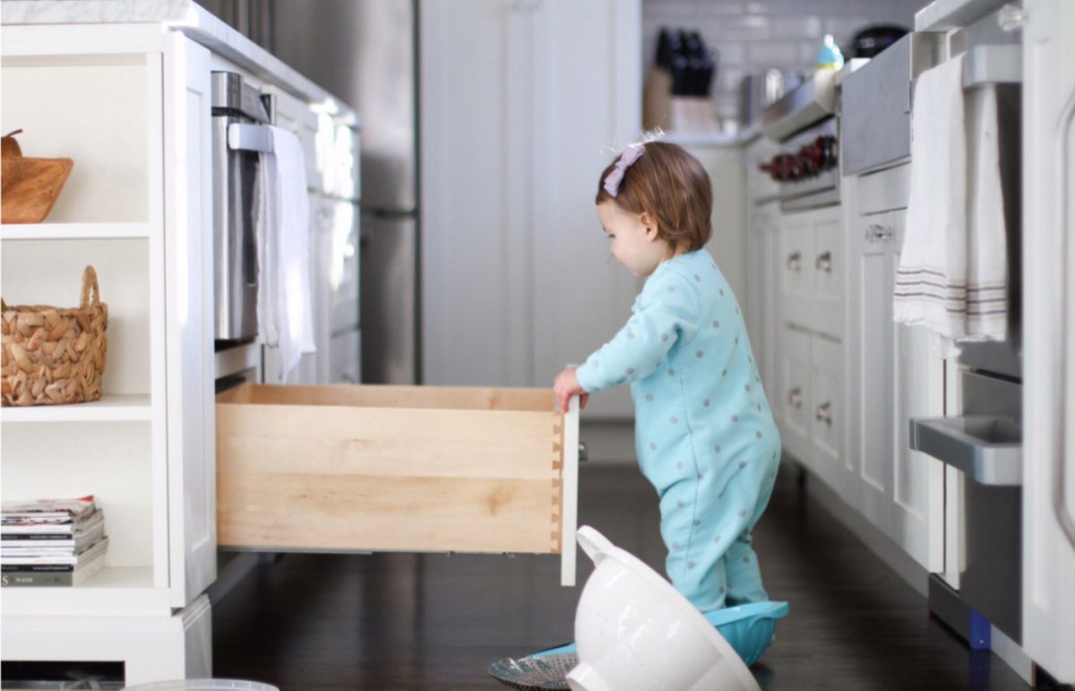 A baby in a blue onsie opening a lower cabinet drawer.