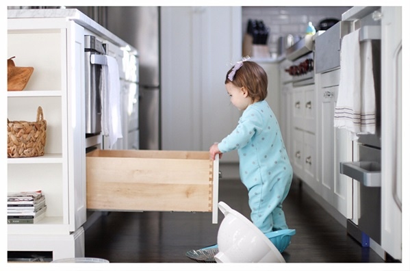 White shaker kitchen and island with toddler girl pulling out cabinet drawer and strainer.