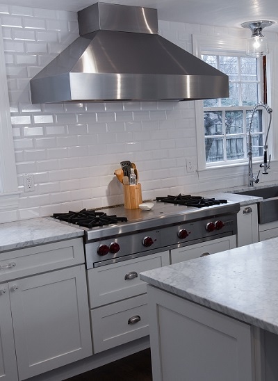 stainless cooktop and hood on wall of glass subway tile with white shaker cabinets