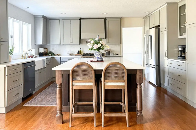 Large Farmhouse-style kitchen with light gray shaker cabinets and a dark wood center island and wicker counter stools and white marble-like quartz countertops.